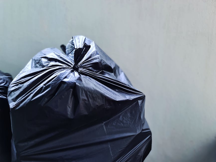 Close-up view of two black plastic rubbish bags filled with waste, stacked and tied at the top, placed against a plain, light-colored wall. The bags are made of shiny, crinkled plastic material, with 